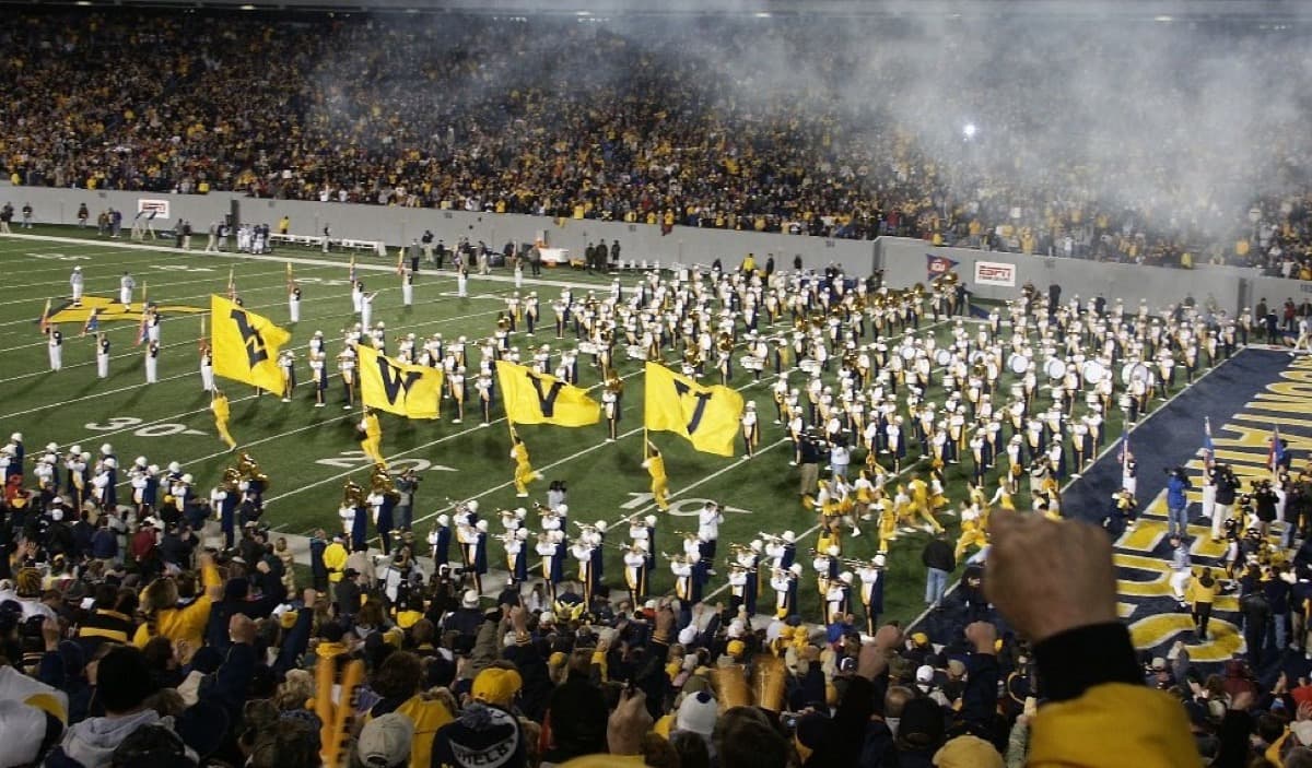 The West Virginia football team running onto Mountaineer Field through smoke and a crowd in gold.